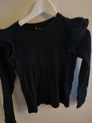 Svart ribbad topp med volangärmar Zara - Svart långärmad topp från Zara med ribbad struktur och volangdetaljer på axlarna. Snygg och trendig modell som ger en cool siluett. Perfekt att styla med jeans eller kjol för en uppdaterad look.