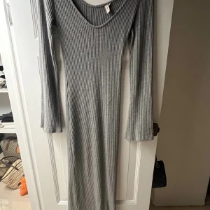 Grå ribbad långklänning - Säljer en lång, ribbad klänning i grått med v-ringning och långa ärmar. Klänningen har en figurnära passform och är tillverkad i ett mjukt stickat material som känns både bekvämt och stilrent. Har en tråd som gått upp