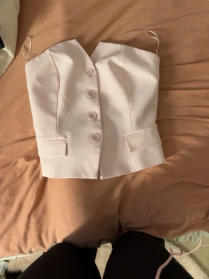 Ljusrosa croppad topp med knappar - Söt ljusrosa croppad kostym topp med knappar framtill. Toppen har volangdetalj baktill och är figursydd med dekorativa fickor framtill. 