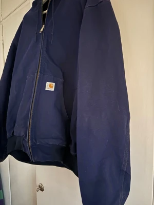 Vintage Carhartt 1993 work jacket med huva. Made in USA - Säljer en mörkblå work jacket  från Carhartt i Canvas med huva. Jackan har en stor ficka framtill med Carhartt-logga och ribbade muddar vid ärmslut och nederkant. XL men sitter snarare som en L. Made in USA och köpt 1993 i Stockholm. Snygg patina och slitningar men inga hål. Foder och muddar i bra skick. Modell 184 cm lång.