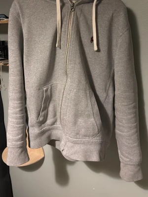 Ralph lauren zip  - PASSAR S. Snygg grå zip hoodie från Polo Ralph Lauren med vit dragsko i huvan och klassisk känguruficka framtill. Liten mörkröd logga broderad på bröstet. Tillverkad i mjuk bomullsblandning, perfekt för chill dagar. Ribbstickade muddar vid ärmslut och nederkant.
