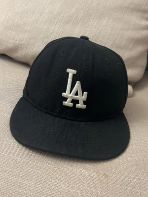 Svart LA-keps från New Era - Säljer en svart keps från New Era med LA-logga broderad i vitt framtill och New Era-logga på sidan. Klassisk rak skärm och justerbar passform. Perfekt för dig som gillar streetwear och sportig stil.