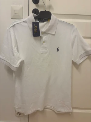 Vit pikétröja från Polo Ralph Lauren - Klassisk vit pikétröja från Polo Ralph Lauren med korta ärmar och blå broderad logga på bröstet. Tröjan har krage och knappar framtill. Perfekt till jeans eller shorts för en clean och stilren look.