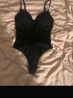 snygg body  - Snygg svart body med smala, justerbara axelband och v-ringad urringning. Modellen har hög skärning vid benen och är tillverkad i ett stretchigt material som sitter tight mot kroppen. Perfekt att styla under en skjorta eller kavaj.