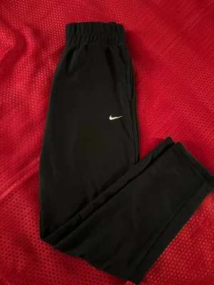 Svarta mjukisbyxor från Nike M  - Svarta mjukisbyxor från Nike med klassisk vit logga på benet. Byxorna har elastisk midja och raka ben, perfekta för chill eller träning. Tillverkade i mjukt material som känns skönt mot huden. Enkel och stilren design som funkar till det mesta.