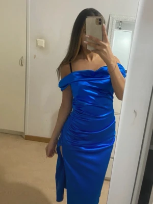 Blå axelbandslös långklänning satin - Snygg blå långklänning i satin med offshoulder-design och slits på sidan. Klänningen har en figurnära passform och draperade detaljer längs sidorna för en extra stylish look.