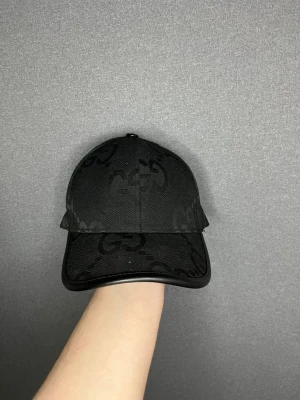 Svart keps från Gucci med GG-mönster - Snygg svart keps från Gucci med det klassiska GG-monogrammet ton-i-ton över hela kepsen. Kepsen har böjd skärm och är tillverkad i ett slitstarkt tygmaterial. Perfekt accessoar för en trendig och stilren look.