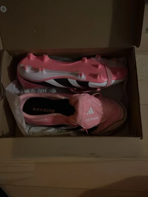 Adidas Beckham rosa fotbollsskor - Säljer ett par snygga Adidas Beckham fotbollsskor i rosa och vitt med svarta detaljer. Skorna har klassisk snörning, dobbar för gräsplan och en stilren design med Beckham-tryck på plösen. Perfekta för dig som vill sticka ut på planen! Ni får såklart buda!!❣️