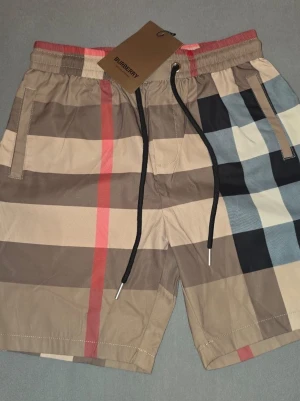 Rutig shorts från Burberry - Säljer ett par klassiska Burberry shorts med det ikoniska rutiga mönstret i beige, svart, rött och vitt. Shortsen har elastisk midja med svart snörning och fickor på sidorna samt en bakficka. Perfekta för sommaren och riktigt snygga till streetwear-stilen. Helt nya burberry shorts med lapp på. Hör av er vid frågor! 