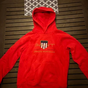 Röd hoodie från GANT med brodyr - Säljer en röd hoodie från GANT med broderad logga och sköld på bröstet. Tröjan har huva, känguruficka och är tillverkad i mjuk bomullsmix. Perfekt för dig som gillar sportig och klassisk stil.