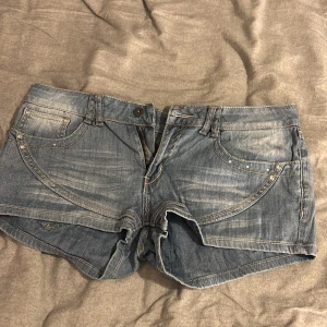 Blå jeansshorts med detaljer, stl 31 - Snygga blå jeansshorts med slitningar och coola sömdetaljer på fickorna. Shortsen har klassisk femficksdesign, dragkedja och knapp framtill samt dekorativa bakfickor med broderi och små nitar. Perfekta för sommardagar och festivalhäng.