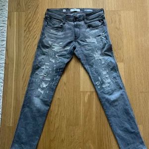 Replay Anbass grå slim jeans, stl 30/32 - Snygga grå Replay Anbass jeans i slim fit med riktigt feta slitningar. Jeansen är tillverkade i ekologisk bomull och har klassisk femficksdesign. Perfekta för dig som gillar en modern och edgy look.