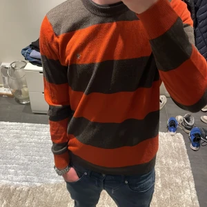 Randig orange och brun tröja från Morris  - Säljer en snygg 100% merinoull tröja från Morris som är randig. Ny pris är ca 1800kr, mitt pris 449kr! Modellen är ca 180cm. Hör av dig vid funderingar!