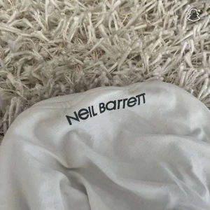 Vit Neil Barrett t-shirt med blixtar - Säljer en vit t-shirt från Neil Barrett med ikoniska svarta blixtar runt halsen. T-shirten har rund halsringning och korta ärmar. Perfekt för dig som vill sticka ut med en cool och edgy stil.