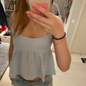 Ljusblå linnetopp med rosett i ryggen - Supersöt ljusblå linnetopp med tunna axelband och volangliknande nederdel från Nelly. Toppen har en snygg öppen rygg med stor knytrosett som detalj. Perfekt för varma dagar och ger en luftig känsla. 