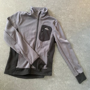 Grå och svart fleecejacka från Protech - Säljer en grå och svart fleecejacka från Protech med hel dragkedja och hög krage. Jackan har en svart bröstficka med dragkedja och två sidofickor. Perfekt för lager-på-lager och chill dagar ute.