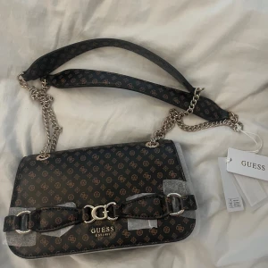 Brun axelväska från Guess med kedja - Brun axelväska från Guess med bruna logomönster över hela väskan. Väskan har silverfärgade kedjedetaljer och ett dekorativt GG-spänne framtill. Justerbar axelrem i konstläder och metall. Helt oanvänd med etikett