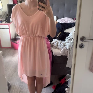 Ljusrosa omlottklänning med resår - Superfin ljusrosa klänning med omlottkänsla och resår i midjan. Klänningen har kortare framstycke och längre bak, vilket ger en snygg och flowy look. Tillverkad i ett tunt, lätt material med mjuk känsla. Perfekt för sommarens alla tillfällen!