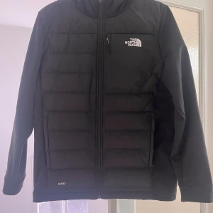Svart pufferjacka från The North Face - Svart pufferjacka från The North Face med huva och diskret logga på bröstet. Jackan har quiltad framsida, dragkedja hela vägen och en bröstficka med zip. Perfekt för kalla dagar och har en clean, sportig look.