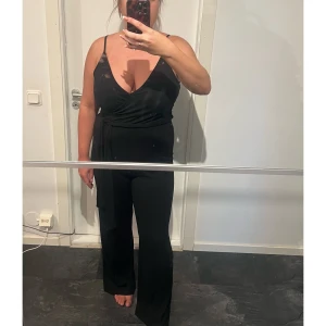 Svart omlott jumpsuit med bälte - Snygg svart jumpsuit med omlott upptill, smala axelband och v-ringning. Byxorna är vida och har ett medföljande bälte i midjan. Materialet är mjukt och har en lätt ribbad struktur som ger en cool look.