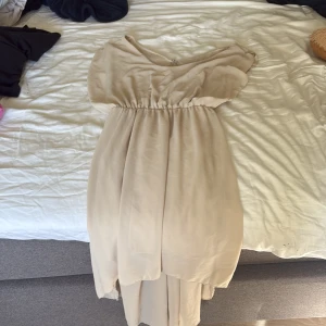 Beige långklänning med omlott och volang - Säljer en elegant beige långklänning i ett lätt och flowy material. Klänningen har omlott-detalj upptill, v-ringning och volang vid axlarna. Midjan är markerad med resår och baktill finns en liten guldfärgad knapp. Perfekt för dig som gillar en romantisk och stilren look.