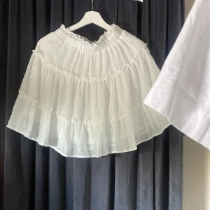 Vit volangkjol från SHEIN - Superfin vit kortkjol från SHEIN med flera lager volanger och resår i midjan. Kjolen är luftig och har en lätt, krinklad struktur som ger en cool och avslappnad vibe. Perfekt att styla med en crop top eller oversized hoodie.