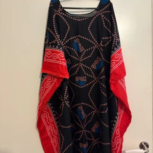 Somalibaati-Svart och röd långklänning med mönster - Unik långklänning i svart med röda kanter och coola prickiga mönster i blått och vitt. Klänningen har en lös passform och vida ärmar, perfekt för dig som gillar statement-plagg. Materialet känns lätt och flowy, och mönstret ger en riktigt snygg boho-vibe.(one seize)