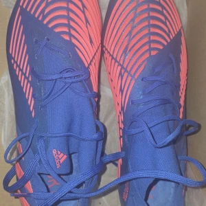 Adidas Predator Edge.1 SG fotbollsskor blå/röd - Säljer ett par Adidas Predator Edge fotbollsskor i blått med röda detaljer. Skorna har snörning, mönstrad ovandel och robust sula med dobbar för bästa grepp på planen. Tillverkade i syntetmaterial och levereras med tillhörande meshpåse. SG = Skruvdobb 