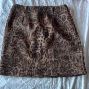 Leopardmönstrad kortkjol med dragkedja - Snygg kortkjol med leopardmönster i bruna och svarta toner. Kjolen har en rak passform och en synlig dragkedja bak som detalj. Perfekt för dig som vill sticka ut med ett djurmönstrat plagg. Materialet känns mjukt och stadigt.