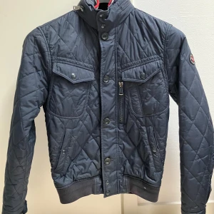 Mörkblå quiltad jacka Polo Ralph Lauren - Quiltad mörkblå jacka från Polo Ralph Lauren med ribbade muddar, hög krage och flera fickor med både knappar och dragkedja. Snygg badge på ärmen och coolt rutmönster över hela jackan. Perfekt för dig som gillar stilren och sportig look..en riktig snygg klassiker som är perfekt i höst och vår. Jackan är i jättebra skick och har använts bara i en halv säsong. XS ca 163-174cm. Finns på olika sajter så först till kvarn! Nypris ca 3000