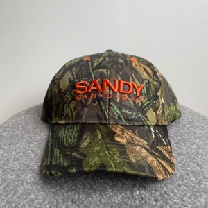 Camo keps från Sandy Goods - Snygg keps från Sandy Goods med camouflagemönster i grönt, brunt och svart. Broderad logga i orange framtill och justerbar kardborre bak. Helt ny, aldrig använd 