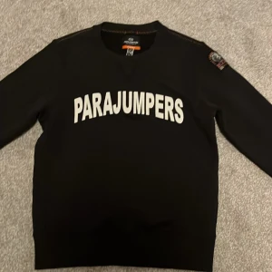 Svart fleece tröja från Parajumpers - Säljer en svart fleece tröja från Parajumpers med vit text på framsidan. Tröjan har långa ärmar och en broderad patch på ärmen. Perfekt för kyliga dagar. Har aldrig kommit till användning så skicket är 10/10