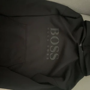 Svart hoodie från Hugo Boss - Svart hoodie från Hugo Boss med stor BOSS-logga på bröstet. Klassisk huva och magficka framtill. Mjuk insida och stilren design, perfekt för dig som gillar streetwear och snygga märkesplagg.