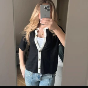 Svart kofta från Massimo Dutti XS - Svart kofta från Massimo Dutti i tunn stickad kvalitet med kontrasterande vit kant och snygga guldknappar framtill. Kortärmad modell med rak passform, perfekt till jeans eller kjol. Klassisk och stilren look som funkar året runt.