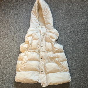 Vit dunväst med huva och snörning - Säljer en lång, vit dunväst med stor huva och dragsko i midjan. Västen har tryckknappar framtill och två fickor på sidorna. Perfekt för lager-på-lager och ger en cool, puffer-inspirerad look.