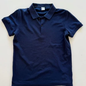 Marinblå pikétröja från LINDEX - Klassisk marinblå pikétröja från LINDEX med krage och två knappar framtill. T-shirten är kortärmad och tillverkad i mjuk bomull, perfekt för en clean och stilren look.