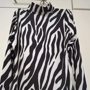 Zebramönstrad polotopp från Nama - Säljer en svartvit zebramönstrad polotopp från Nama i storlek M. Toppen har lång ärm, hög krage och dragkedja i ryggen. Materialet är stretchigt och känns mjukt mot huden. Perfekt för dig som vill sticka ut med ett djurmönster.