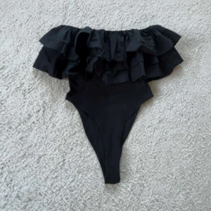 Svart volangbody från Zara - Supersnygg svart body från Zara med bred volang upptill som ger en trendig och cool look. Perfekt för  när du vill sticka ut lite extra. Modellen är axelbandslös och har en hög benskärning.