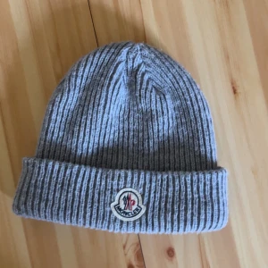 Grå ribbstickad mössa från Moncler - Snygg grå ribbstickad mössa från Moncler med uppvikt kant och klassisk Moncler-logga framtill. Perfekt för kalla dagar och ger en clean look till din outfit. Tillverkad i mjukt material som sitter skönt på huvudet. Pris kan diskuteras