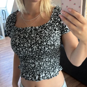 Svartvit blommig croppad blus - Säljer en svartvit blommig croppad blus med korta puffärmar och smockad midja. Toppen har volangkant nertill och kan bäras både på axlarna och offshoulder. Perfekt för sommarens alla tillfällen! Väldigt skönt material också, använd minst två gånger!