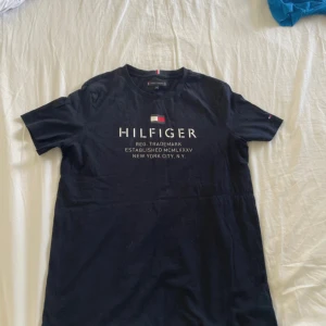 Mörkblå t-shirtklänning Tommy Hilfiger - Snygg mörkblå t-shirtklänning från Tommy Hilfiger med klassisk logga och tryck framtill. Rak passform, rund halsringning och korta ärmar. Perfekt för en chill och stilren look. Tillverkad i mjuk bomull för maximal komfort.