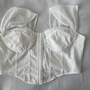 Vit korsettopp med transparenta axelband - Supersnygg vit korsettopp med markerade kupor och transparenta, puffiga axelband i organza. Toppen har en figurnära passform och dekorativa sömmar framtill som ger en cool och trendig look. Perfekt att styla med jeans eller kjol för en chic vibe.