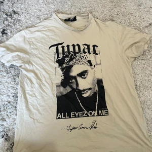 Beige Tupac t-shirt från Bershka XS - Snygg beige t-shirt från Bershka med stort Tupac-tryck och texten 'All Eyez On Me' framtill. Baksidan har låtlista från albumet. Klassisk passform och rund hals. Perfekt för dig som gillar streetstyle och hiphop-vibe. Den är stor i storlek! 