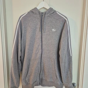 Grå hoodie från Adidas med dragkedja - Grå hoodie från Adidas med klassiska vita ränder längs ärmarna och Adidas-logga på bröstet. Tröjan har huva, hel dragkedja och två stora fickor framtill. Tillverkad i mjuk bomullsblandning, perfekt för chill eller träning.