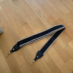 Svart och beige axelrem till väska - Snygg bred axelrem i svart och beige med geometriskt mönster. Remmen har guldfärgade metalldetaljer och är justerbar. Perfekt för att ge din väska en ny look eller byta ut en gammal rem.