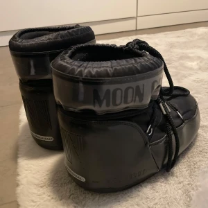 Svarta Moon Boot Original - Säljer ett par svarta Moon Boot Original med klassisk rund form och bred platt sula. Skorna har snörning framtill och är tillverkade i syntetmaterial med vadderad insida för extra värme. Perfekta för vintern och riktigt sköna att ha på sig när det är kallt. 