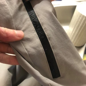 Gråa Emporio Armani EA7 byxor - Snygga gråa byxor från Emporio Armani EA7 med svarta detaljer och dragkedjor. Byxorna har elastisk midja och logga på benet. Perfekta för en sportig och stilren look. Materialet känns mjukt och bekvämt mot huden.