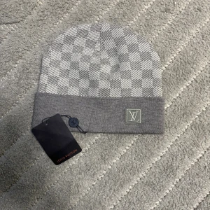 Grå rutig mössa från Louis Vuitton - Snygg grå mössa från Louis Vuitton med klassiskt rutigt mönster och vikt kant. LV-logga framtill i broderad patch. Mjuk och skön stickad kvalitet, perfekt för kyliga dagar. En stilren accessoar som lyfter din look.