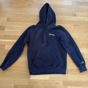 Svart Champion hoodie med huva - Säljer en svart hoodie från Champion i storlek S. Tröjan har klassisk huva med snörning, magficka och broderad Champion-logga på bröstet och ärmen. Smal passform. 
