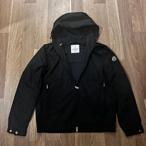 Moncler jacka  - Svart vindjacka från Moncler med huva och dragkedja framtill. Jackan har två sidofickor med dragkedja, tryckknappar vid ärmslut och Moncler-logga på ena ärmen. Lätt och smidig modell i syntetmaterial, perfekt för blåsiga dagar.